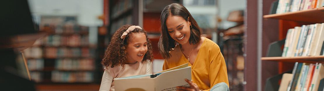 A teacher sits in a library with a young student reading a book